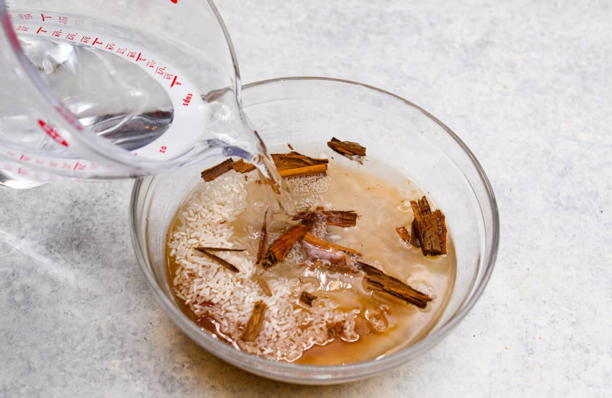 Elevate the flavors of traditional horchata with this strawberry horchata recipe! It's a creamy drink soaked in rice water and cinnamon sticks, then sweetened with a strawberry puree. Everyone loves the strawberry version of this rich, milky Mexican drink! Process photo of water pouring into a bowl of rice and cinnamon sticks.