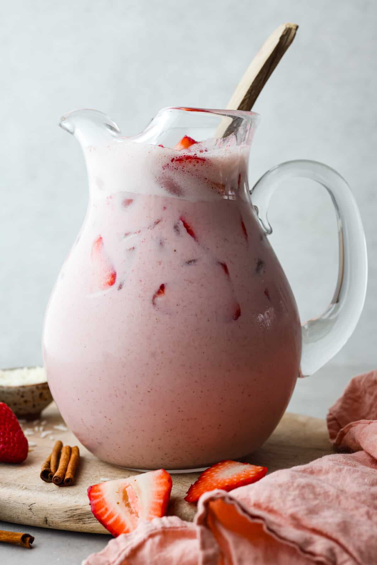 Elevate the flavors of traditional horchata with this strawberry horchata recipe! It's a creamy drink soaked in rice water and cinnamon sticks, then sweetened with a strawberry puree. Everyone loves the strawberry version of this rich, milky Mexican drink! Close view of strawberry horchata in a glass pitcher with a large gold spoon for stirring.