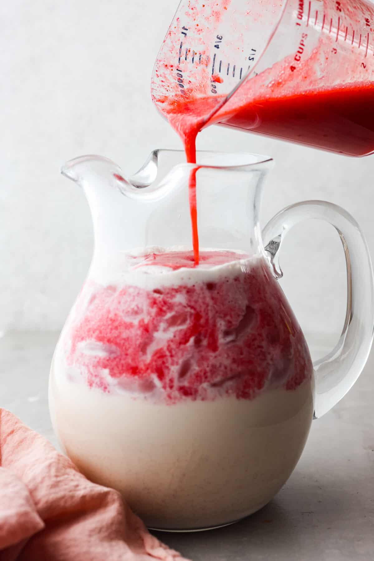 Elevate the flavors of traditional horchata with this strawberry horchata recipe! It's a creamy drink soaked in rice water and cinnamon sticks, then sweetened with a strawberry puree. Everyone loves the strawberry version of this rich, milky Mexican drink! Close view of a large glass pitcher full of horchata with the strawberry puree pouring in.