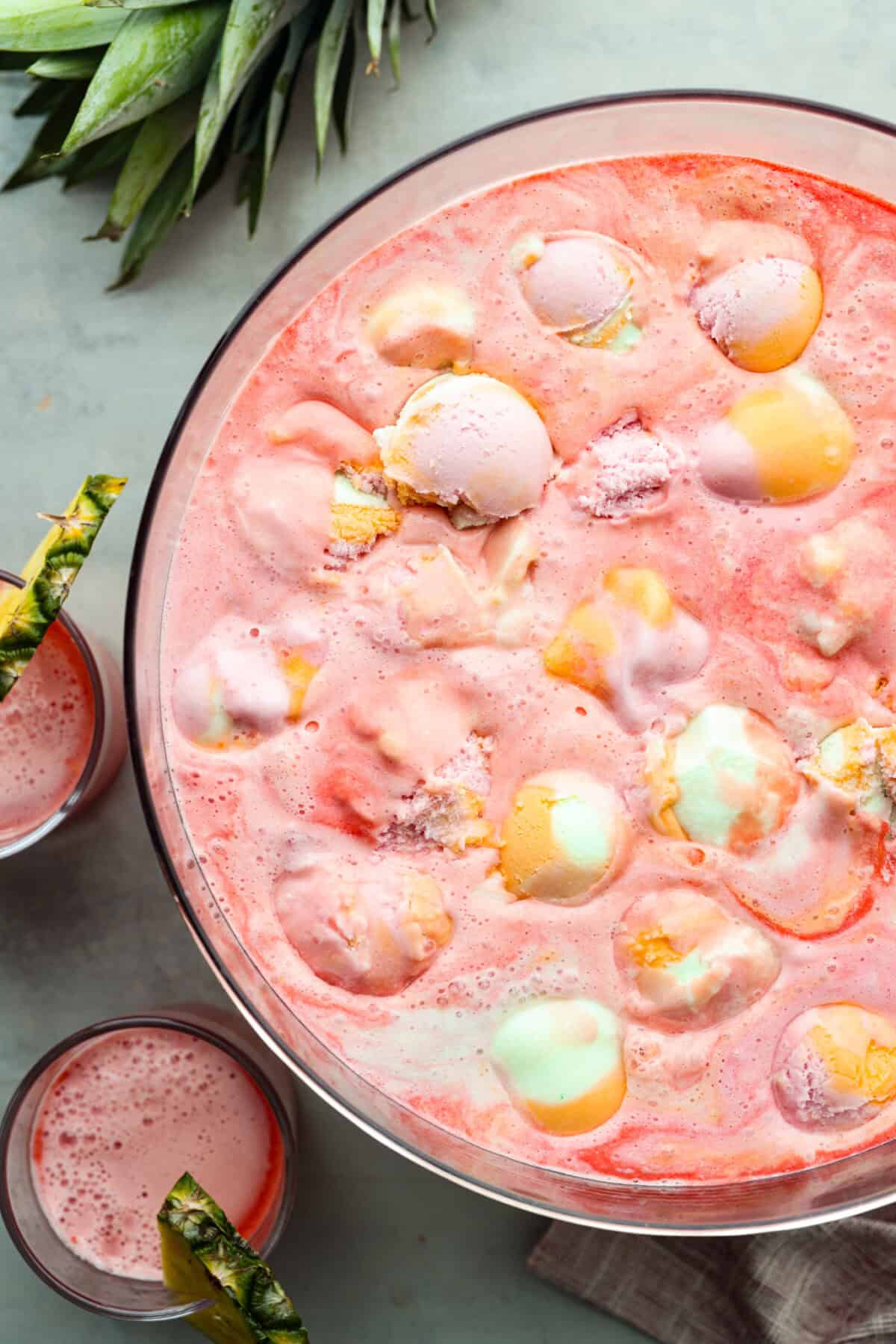 This bright sherbet punch is perfect for serving on any occasion! It has just the right balance of sweetness, zest, and refreshing coolness and you won't be able to resist having a second glass. Overhead shot of a large punch bowl filled with sherbet punch next to smaller cups with the drink in them and a pineapple wedge on the rim of the glass.