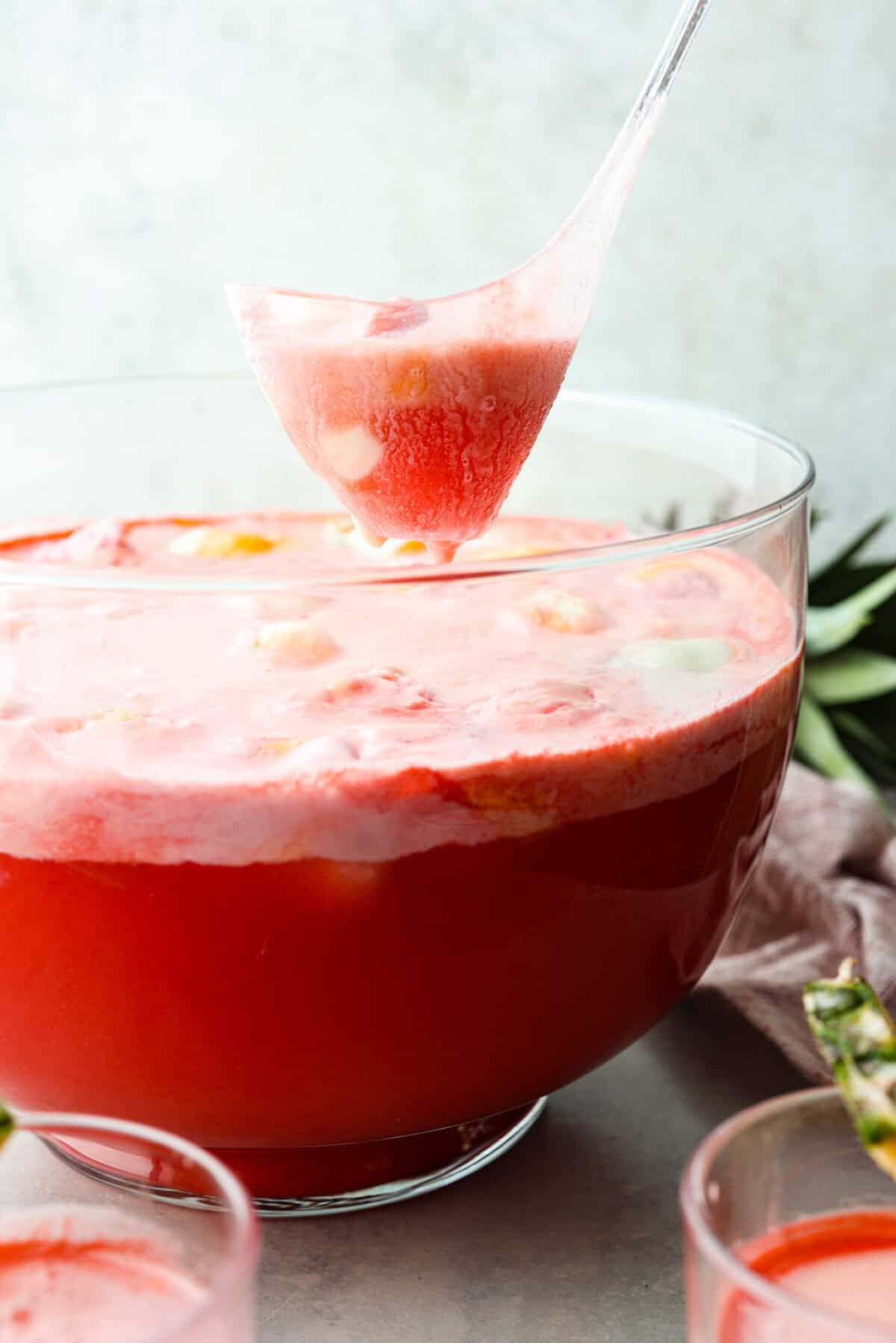 This bright sherbet punch is the perfect addition to any BBQ or gathering. Your guests will keep coming back for more and beg you for the recipe! It has just the right balance of sweetness, zest, and refreshing coolness, you'll be sure to beat the heat on a hot summer day. Side view of a large bowl filled with bright red punch that is topped with rainbow sherbet that has foam and big chunks of the sherbet.