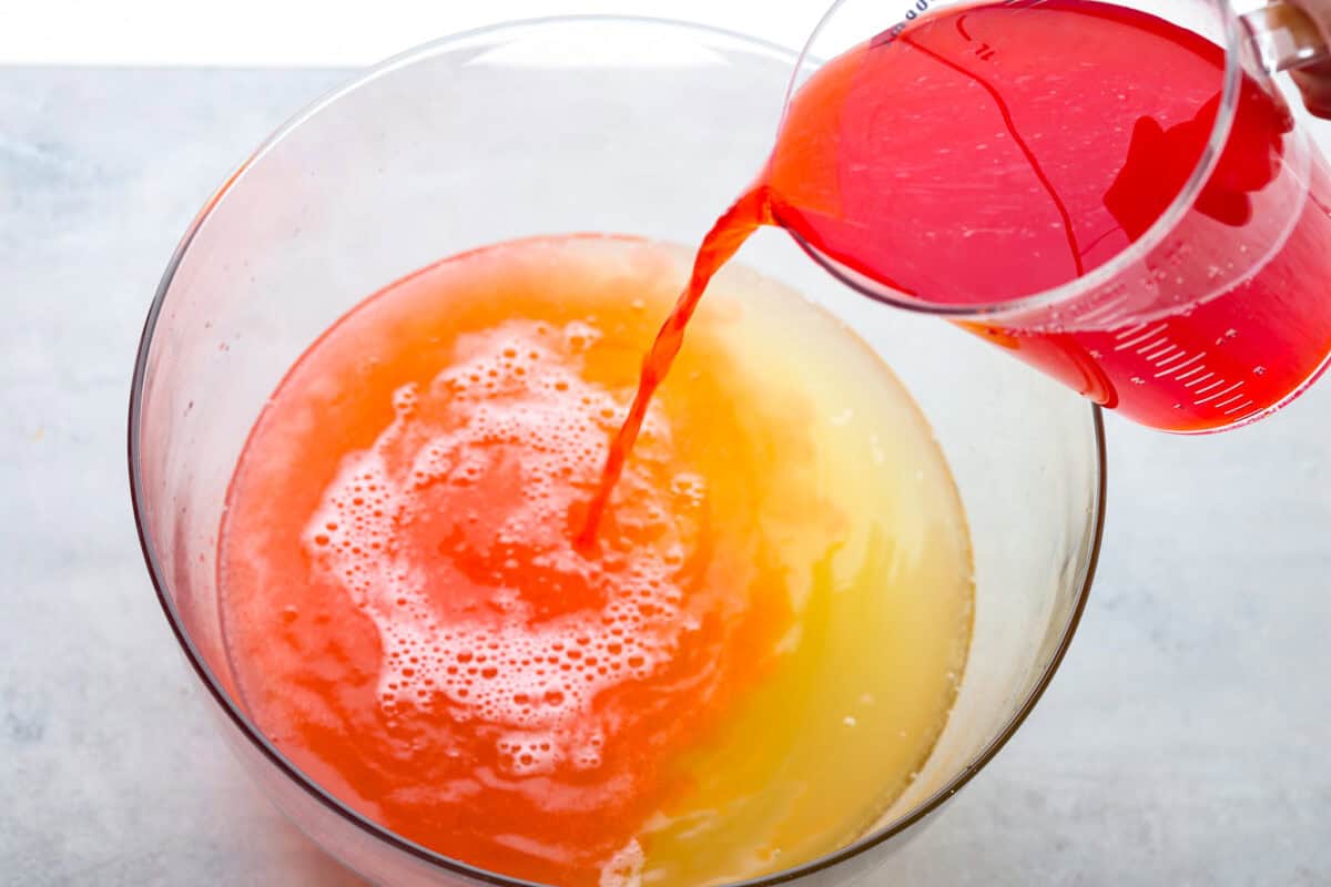 This bright sherbet punch is the perfect addition to any BBQ or gathering. Your guests will keep coming back for more and beg you for the recipe! It has just the right balance of sweetness, zest, and refreshing coolness, you'll be sure to beat the heat on a hot summer day. This is an overhead shot of a large bowl that has Sprite and pineapple juice in it. There is also a large measuring cup tipping its contents of Hawaiian Punch into the large bowl.