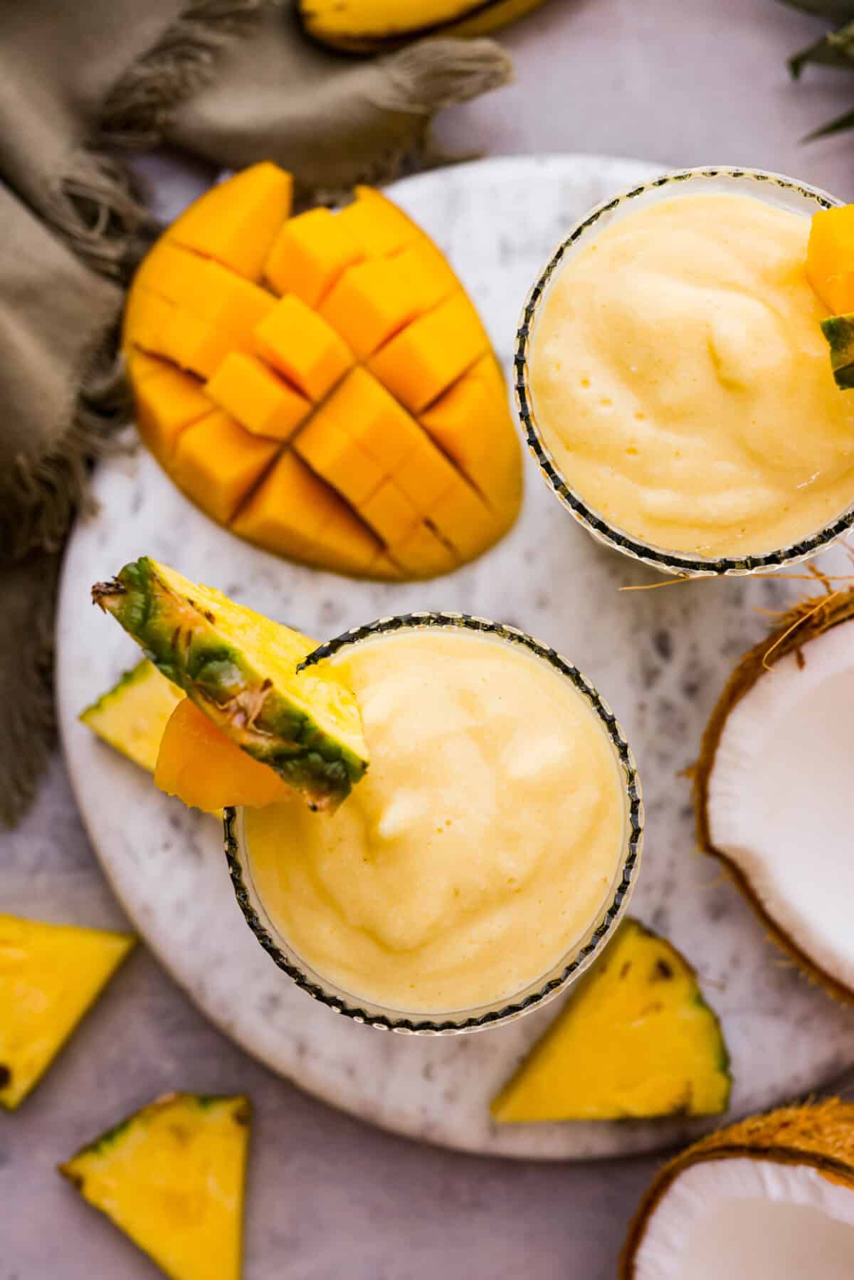 This refreshing tropical smoothie is bursting with tropical fruit flavors, making it the perfect treat for a hot day. Sip it, and you'll feel like you're lounging on the beach, enjoying a drink freshly served by the cabana boy! Overhead shot looking down at the top of two glasses next to pineapple wedges, cut up mango and half of a coconut.