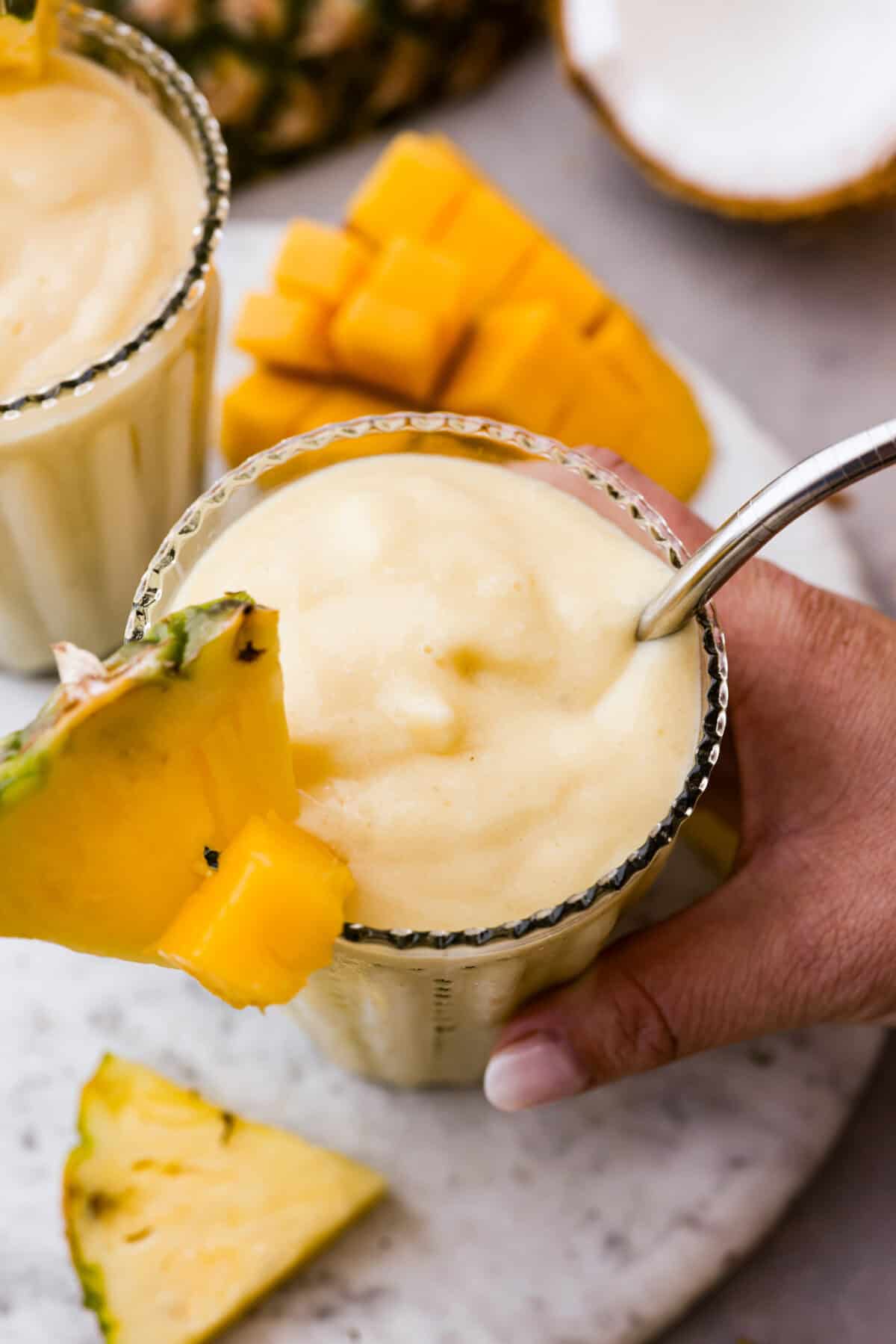 This refreshing tropical smoothie is bursting with tropical fruit flavors, making it the perfect treat for a hot day. Sip it, and you'll feel like you're lounging on the beach, enjoying a drink freshly served by the cabana boy! Angle shot of someone picking up the glass of tropical smoothie with wedge of pineapple in it.