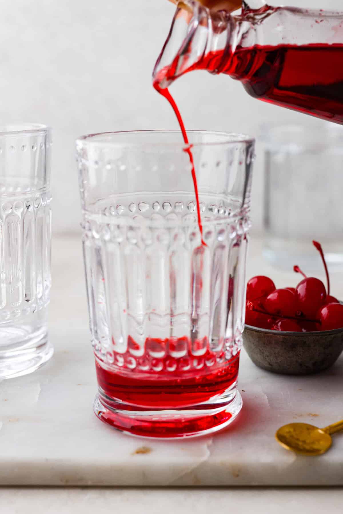 The Shirley Temple is a classic, non-alcoholic drink that's as fun as it is refreshing! Made with just a few simple ingredients, its sweet, bubbly flavor and signature cherry garnish make it a favorite for both kids and adults! Side shot of someone pouring grenadine into the bottom of a glass.