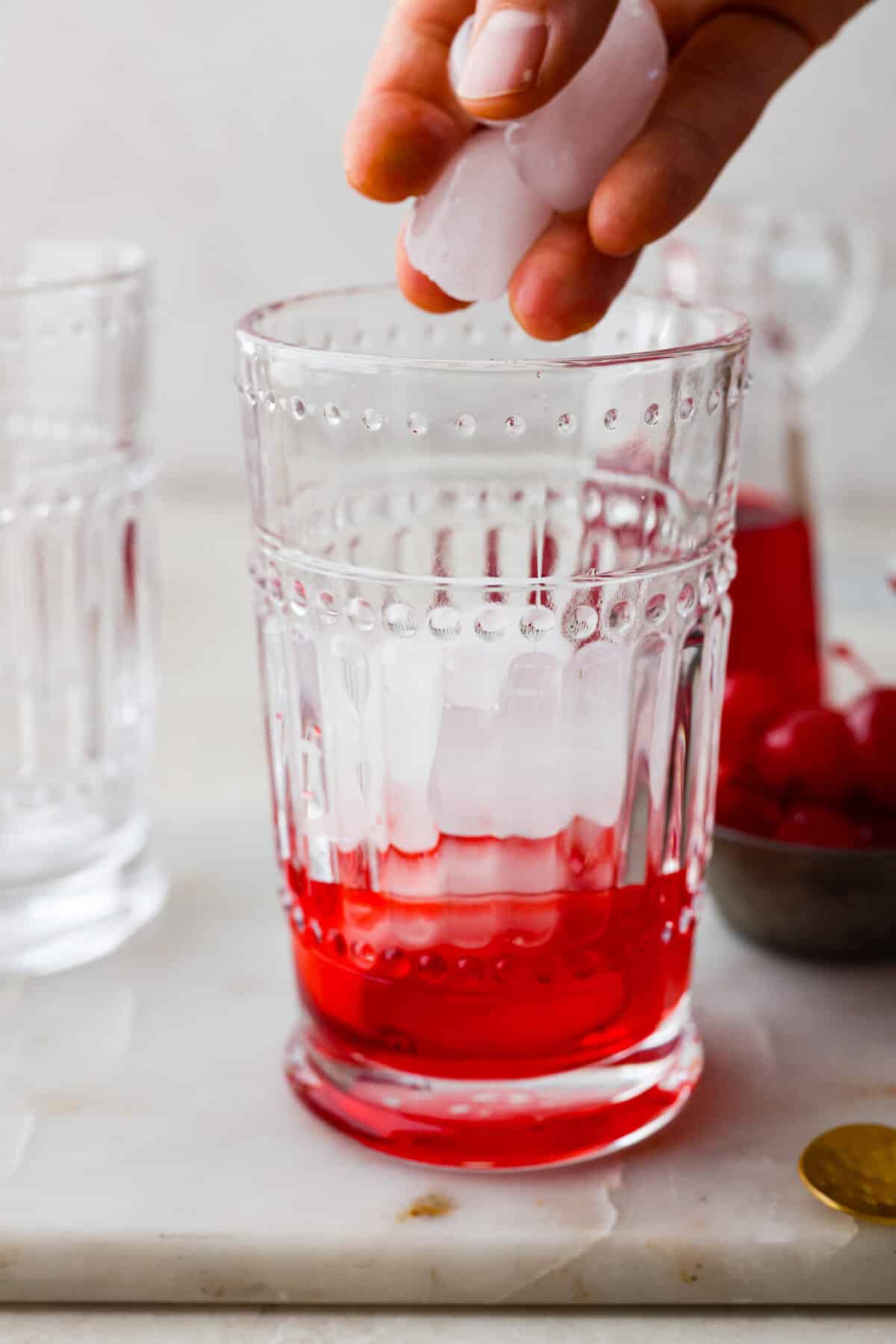 The Shirley Temple is a classic, non-alcoholic drink that's as fun as it is refreshing! Made with just a few simple ingredients, its sweet, bubbly flavor and signature cherry garnish make it a favorite for both kids and adults! Side shot of someone putting ice into the glass with grenadine.
