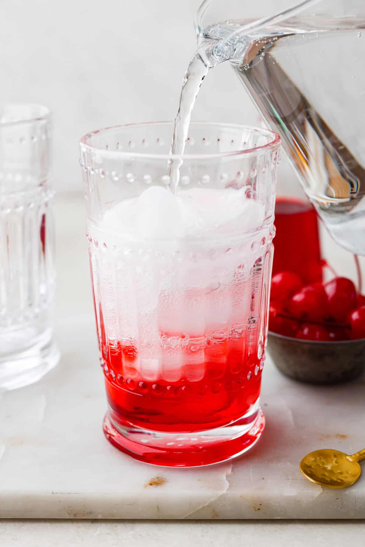 The Shirley Temple is a classic, non-alcoholic drink that's as fun as it is refreshing! Made with just a few simple ingredients, its sweet, bubbly flavor and signature cherry garnish make it a favorite for both kids and adults! Side shot of someone pouring soda into the ice and grenadine to complete their Shirley Temple.