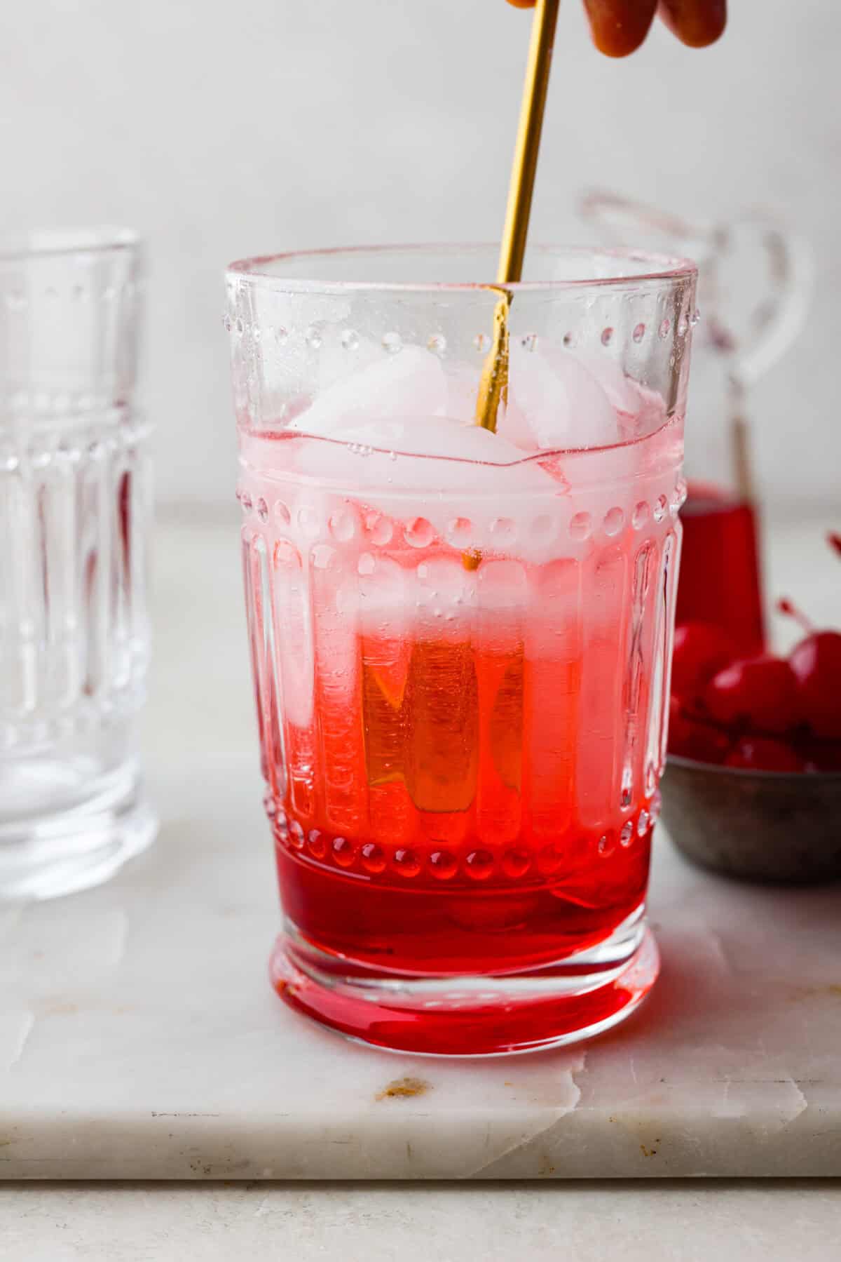 The Shirley Temple is a classic, non-alcoholic drink that's as fun as it is refreshing! Made with just a few simple ingredients, its sweet, bubbly flavor and signature cherry garnish make it a favorite for both kids and adults! Side shot of someone stirring the Shirley Temple.