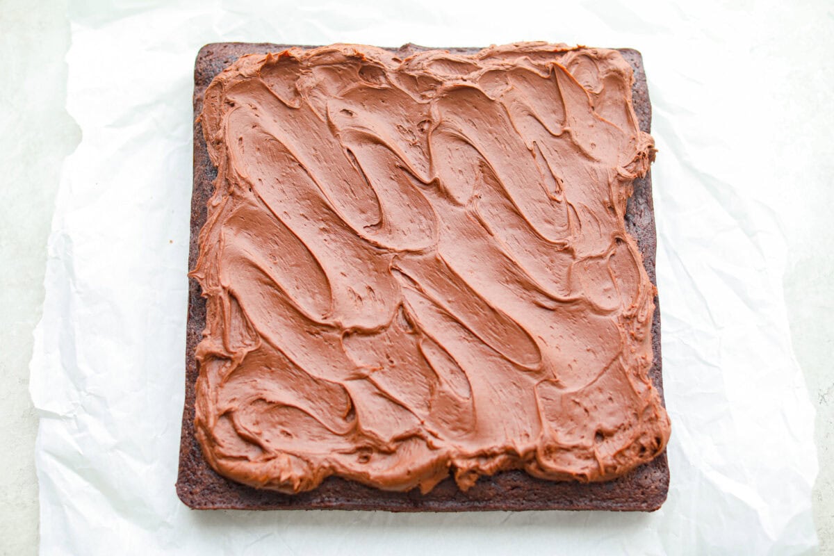 Rich, fudgy, and packed with deep chocolate flavor, Guinness Brownies are a must-try for any dessert lover! The stout beer enhances the cocoa, adding a subtle, malty depth of flavor that makes these brownies rich and irresistibly delicious. Overhead shot of baked and frosted Guinness brownies on a piece of parchment paper.